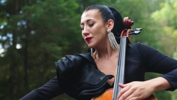 具有绿色自然背景的迷人的女音乐家 华丽的女人戴着华丽的耳环 穿着华丽的衣服 在绿色的森林里弹奏大提琴 — 图库视频影像