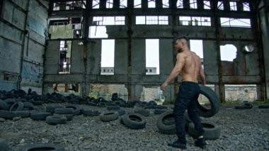 Tişörtsüz adam mahvolmuş bitkide eğitim görüyor. Terk edilmiş yerde spor yaparken eski araba lastiklerini atan güçlü bir atlet. Çapraz fitness rutini.