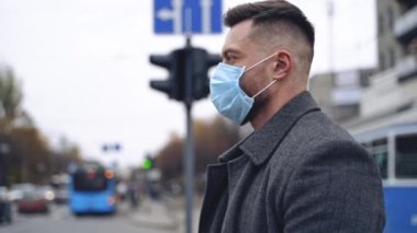 Dışarıdaki maskeli erkek yüzü profili. Koruyucu tıbbi maskeli genç adam şehir ulaşımının yanında duruyor. Salgın Koronavirüs. Yavaş çekim.