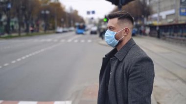 Şehir taşıma geçmişinde maskeli bir adam var. Sıcak palto ve koruyucu sağlık maskesi takmış bir adamın yol kenarında duruşu. Ağır çekim. Virüs koruması.