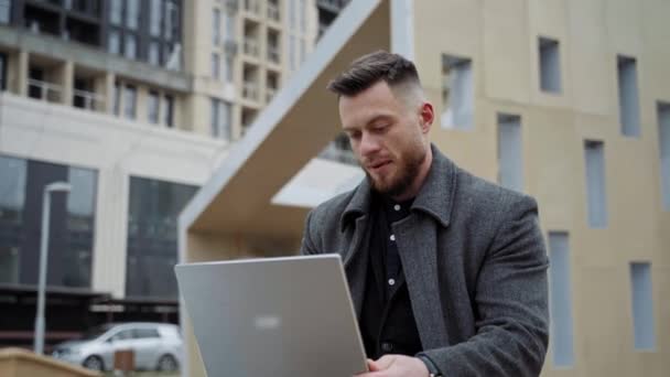 年轻人在新鲜空气上带着笔记本电脑 一个穿着外套的英俊的年轻人坐在街上 在城市里为一个现代化的装置工作 远程工作 — 图库视频影像