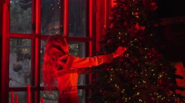 Kadın Noel ağacı süslüyor. Güzel bir kız yeni yıl ağacını süslüyor ve pencereden bakıyor. Güzel noel sahnesi. Kırmızı ışık.