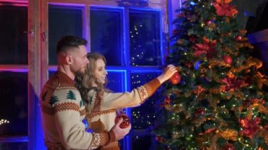 Mutlu çift Noel ağacını süslüyor. Güzel bir kadın Noel toplarını kocasıyla birlikte pencere arkasına asar. Yeni yıl kutlaması.