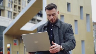 Laptoplu yakışıklı bir adamın portresi. Palto giyen genç sakallı iş adamı, şehir arka planında kablosuz bir cihaz üzerinde çalışıyor..
