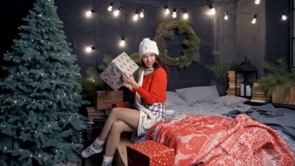 ギフトボックス付きのポジティブな若い女性 編まれた帽子とスカーフの魅力的な少女は 大きなプレゼントとクリスマスツリーの近くのベッドに座ってそれを幸せに投げます ハッピーニューイヤー — ストック動画