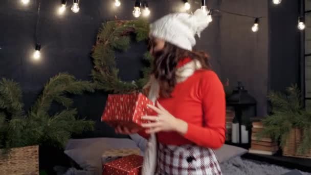 ベッドの上のハッピーガールはギフトボックスを投げます クリスマスプレゼントを保持し 自宅で楽しむニットキャップで美しい若い女性 新しい年 — ストック動画