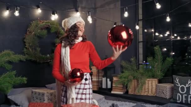大きな赤いクリスマスボールを持つ美しい女性 白いニットのスカーフと帽子の幸せな若い女性は 新年にきれいに装飾された部屋でクリスマスの問題を抱えています — ストック動画
