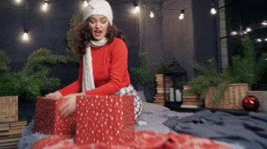 Evde Noel hediyeleri taşıyan mutlu bir kadın. Örgü şapkalı ve atkılı neşeli kız yatakta oturuyor ve Noel süslemelerine hediyeler atıyor..