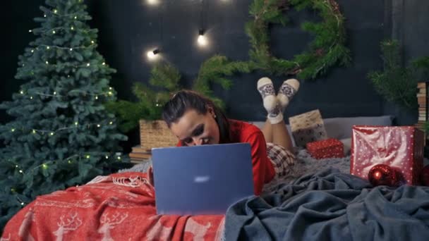 ベッドの上にラップトップを持っている魅力的な少女 美しい若い女性は 新しい年の木の近くのプレゼントとベッドに横たわっている間 ノートパソコンを見ています — ストック動画
