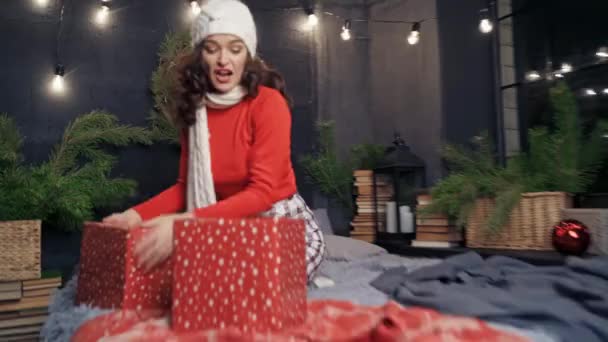 自宅でクリスマスプレゼントを贈るハッピーな女性 編まれた帽子とスカーフで楽しい少女は ベッドの上に座ってクリスマスの装飾の背景にプレゼントを投げます — ストック動画