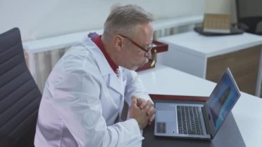 Profesyonel doktor bir hastayla çevrimiçi konuşuyor. Gözlüklü kendine güvenen doktor hastane ofisinde bilgisayarını uzaktan kullanan bir hastaya danışıyor. Uzak uygulama işi.