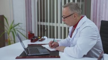 Teletıp sırasında tıp merkezindeki masada oturan bir doktor. Beyaz önlük ve gözlüklü bir tıp uzmanı bir dizüstü bilgisayarla online video görüşmesi yapıyor..