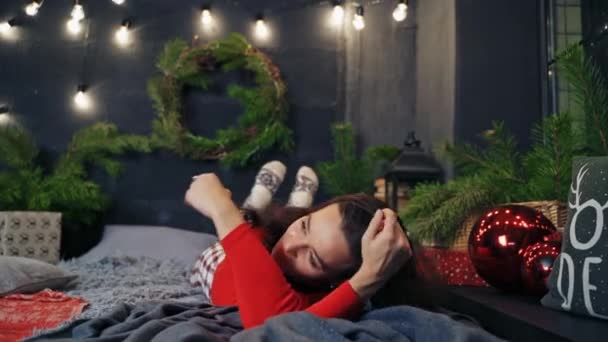 新年のベッドに横たわる陽気な女性 クリスマスの装飾された部屋の背景に喜んでベッドの上に転がるかなり笑顔の若い女性 — ストック動画