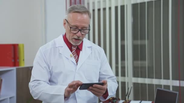 医生戴着眼镜 手里拿着手机 身穿白衣的医学专家与医务室的某个人交谈 — 图库视频影像