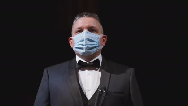 エレガントなスーツとメディカルマスクの男 劇場のステージに立っている保護フェイスマスクの俳優の肖像画 顔からマスクを外す男 コロナウイルスパンデミック — ストック動画