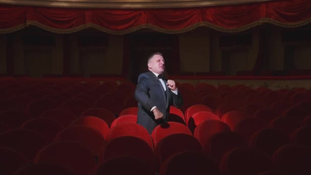 暗い空っぽのシアターホールで幸せな男 大きな講堂で空いている赤いアームチェアの間に立って 一人で楽しく踊るリッチな男 — ストック動画