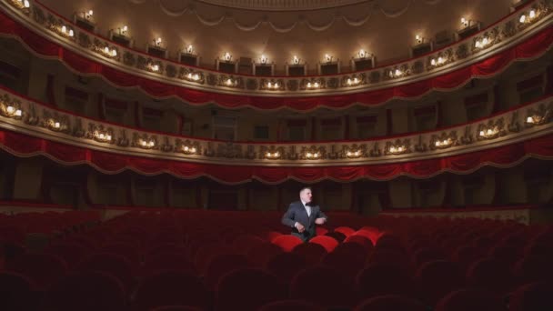 男演员坐在空荡荡的音乐厅里的红色座位上 穿着西装的男人站在漂亮的大空旷的礼堂里 谈论着什么 世界上的禁锢 — 图库视频影像