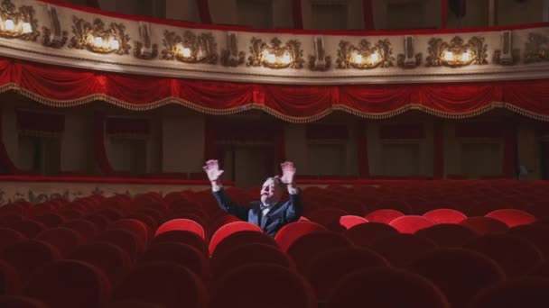 大きな講堂で一人でスーツを着た成功した男 俳優の男は空の赤いアームチェアの間に座ってオペラハウスで歌っています — ストック動画