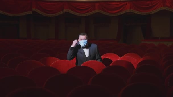 空っぽの劇場にいる男 リッチな男はフェイシャルマスクを脱いで 観客なしで劇場の赤い座席の列の間で何かを言う パンデミックにおける世界危機 — ストック動画