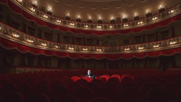 戴面具的观众一个人坐在空荡荡的剧场里 男士们摘下他的面具 在黑暗的大礼堂里 在戏院里一排排满了空的红色椅子中交谈 — 图库视频影像