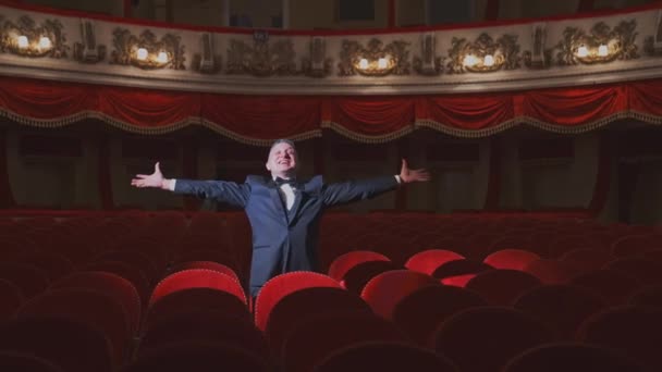 空っぽのシアターホールで幸せな金持ち 劇場の空っぽの椅子の間に一人で立っているスーツで成功したビジネスマンと手を振る — ストック動画