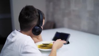 Mutfakta tabletli çocuk. Çocuk yemeğini yiyor ve tabletini izlemekten zevk alıyor.