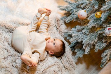 Samimi bir kilimle Noel 'i kutlayan neşeli bir bebek. Halıya uzanmış bir bebek Noel ağacının yanında