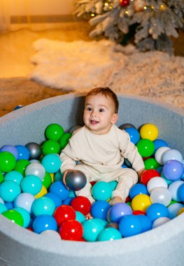 Renkli taşaklarla çevrili neşeli bir bebek, Noel ağacına bakıyor. Arka planda Noel ağacı olan bir bebek top havuzunda oturuyor.