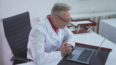 Ofis masasında çalışan erkek doktor. Doktor terapist hasta görüntülü sohbetini klinikteki dizüstü bilgisayarla yapıyor.