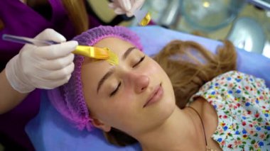 Kozmetik uzmanı, kozmetolojide fırça kullanarak kadının yüzüne altın maske takıyor. Güzellik kliniğindeki güzellik uzmanının cilt temizleme ameliyatındaki genç kız portresi. Yukarıdan görüntüler.