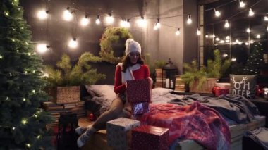 Noel, Yeni Yıl, kış tatili konsepti: Genç mutlu gülümseyen bayan bir sürü hediye tutuyor, süslü püslü evde poz veriyor. Kırmızı kazaklı, beyaz bereli bir manken..