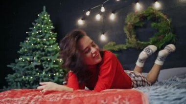 Tatilden önce mutlu bir kız yatakta poz verip yuvarlanıyor. Noel zamanı kırmızı kazaklı güzellik. Görüntü kavramı.