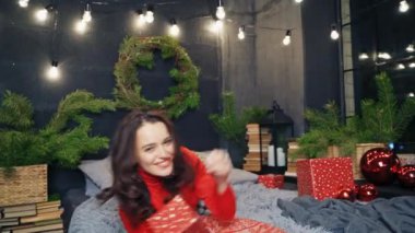 Sıcak ve şık giyinmiş bir kadın dekore edilmiş bir odada yatakta poz veriyor. Kırmızı kazaklı güzel hediye almaktan mutluluk duyar. Eğlence ruhu..