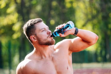 Antrenman sırasında susuz kalan gömleksiz adam. Şişeden su içen üstsüz bir adam.
