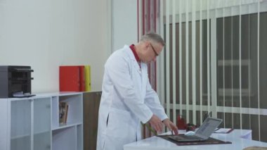 Aydınlık ofisinde dizüstü bilgisayarında yazan deneyimli bir doktor. Modern klinik. Önlüklü doktor. Yakın çekim..