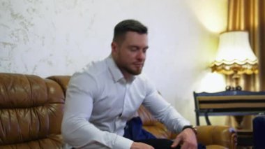Beyaz gömlekli genç yakışıklı bir adam bir otel odasındaki kanepede dinleniyor. İş adamı iş gezisi sırasında bir otele taşındı. Modern otel iç mimarisi.