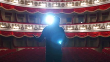 Tiyatroda bir spiker. Bir kişinin parlak yuvarlak ışığı. Siyah takım elbiseli adam konser salonuna gidiyor. Arkadan bak..