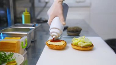 Hamburgerin üzerine ketçap ekliyorum. Mutfakta hamburger hazırlıyorum. Elleri beyaz eldivenlerle pişir ve hamburger yap. Seçici odak.