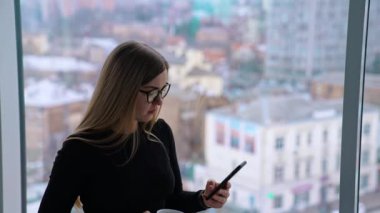 Bir fincan kahveyle güzel bir kadın pencerenin önünde dikilirken dikkatlice akıllı telefona bakıyor.