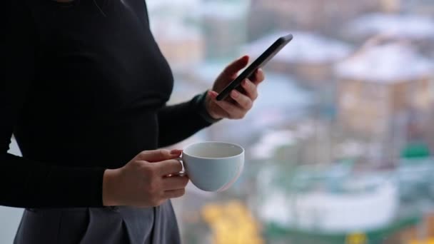 窓のそばに立っている間 思慮深くスマートフォンを閲覧するコーヒーのカップが付いている美しい女性 — ストック動画