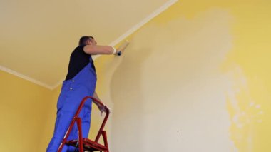 Adam iş yerinde sarı bir duvarı silindirle boyuyor. Profesyonel resim hizmetleri sunuyor..