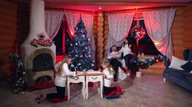 Dört kişilik güzel bir aile Noel için bir odada dinleniyor. Anne ve baba koltukta barışçıl bir şekilde sohbet ediyorlar. Kızlar köknar ağacında resim çiziyor..