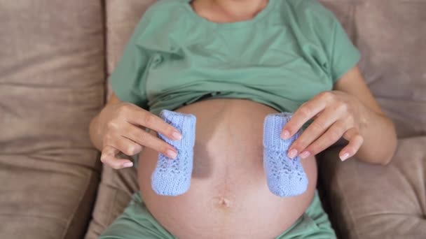 怀孕了 漂亮的女人坐在沙发上 穿着蓝色的小婴儿靴 女人用她那光溜溜的大肚子来回挪动那只小袜子 — 图库视频影像
