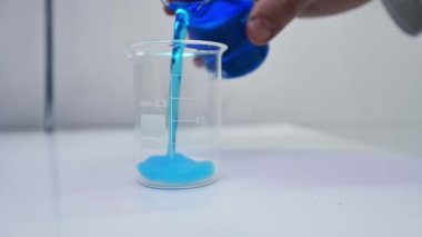 Araştırmacının eli bez dolgusu ile şişeye mavi sıvı döküyor. Mavi sıvı dolguyla karışır ve onunla etkileşime girer. Kapat..