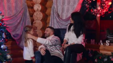 Mutlu ebeveynler rahat koltukta oturur ve neşeyle sohbet ederler. Küçük kızları onlara gelir ve çizimini yapar. Noel ağacı ve dekoratif çelenklerin yanında güzel bir aile.