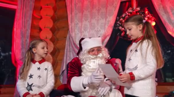 子供たちはサンタの近くに立って 彼に話しかけている 子供たちは写真とクリスマスの夢をサンタクロースと共有しています サンタは休日のために自分の願いについて女の子に尋ねます — ストック動画