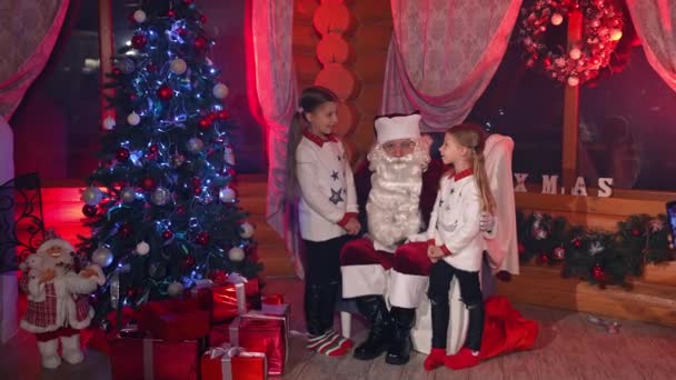 2人の姉妹がサンタのそばに立っていて 彼は椅子に座っている クリスマスにサンタさんに願いを伝える少女たち 火の木が付いている装飾された部屋の子供およびサンタ — ストック動画