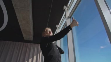Siyah tişörtlü, kameraya bakıp poz veren ve video çeken başarılı bir kendine güvenen kadın. Kadın ofis çalışanı büyük bir ofis penceresinin önünde selfie çekiyor. Aşağıdan görüntüle.