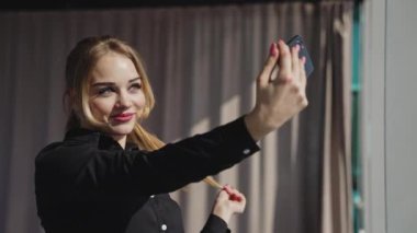 Gülümseyen, genç, çekici, yetişkin bir kadın elinde telefonla selfie çekiyor. Mutlu güzel bayan profesyonel gülümseme, güneşin altında pencerenin yanında tek başına poz verir..