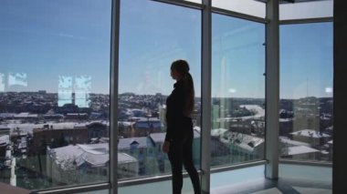 Panoramik pencerelerin önünde duran koyu renk elbiseli genç bir bayan. Güneş ışınları büyük ofis odasına geliyor. Kız pencereden dışarı bakıyor ve şehrin tadını çıkarıyor..
